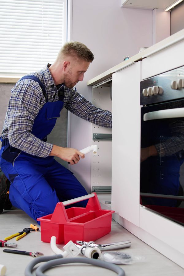A man in blue overalls with a red tool box and extra piping, working under a kitchen sink.