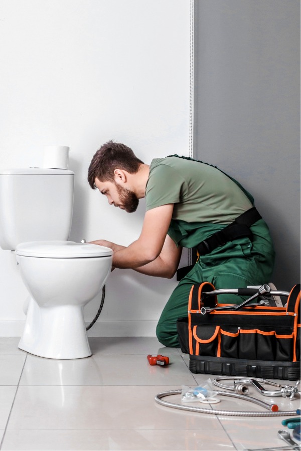 A man bent next to a white toilet with a tool bag, working on the water pipes.