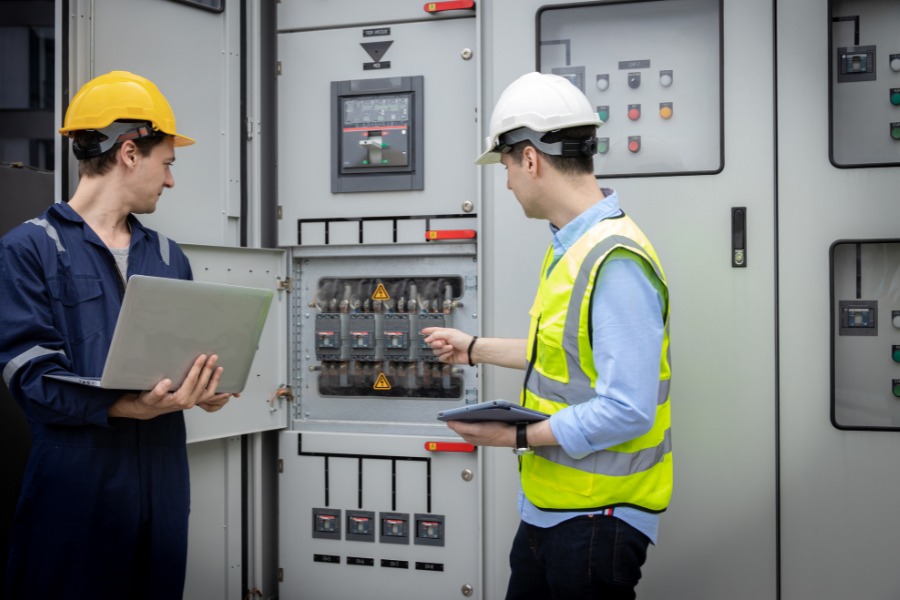Two people around a commercial electrical panel with hard hats and work outfits on.