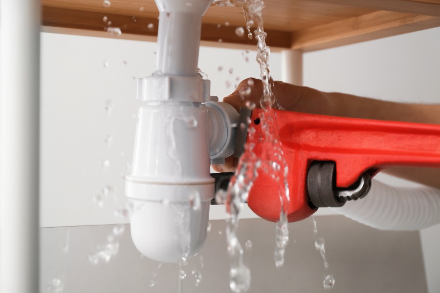 A red wrench twisting a leaking white pipe under a table.
