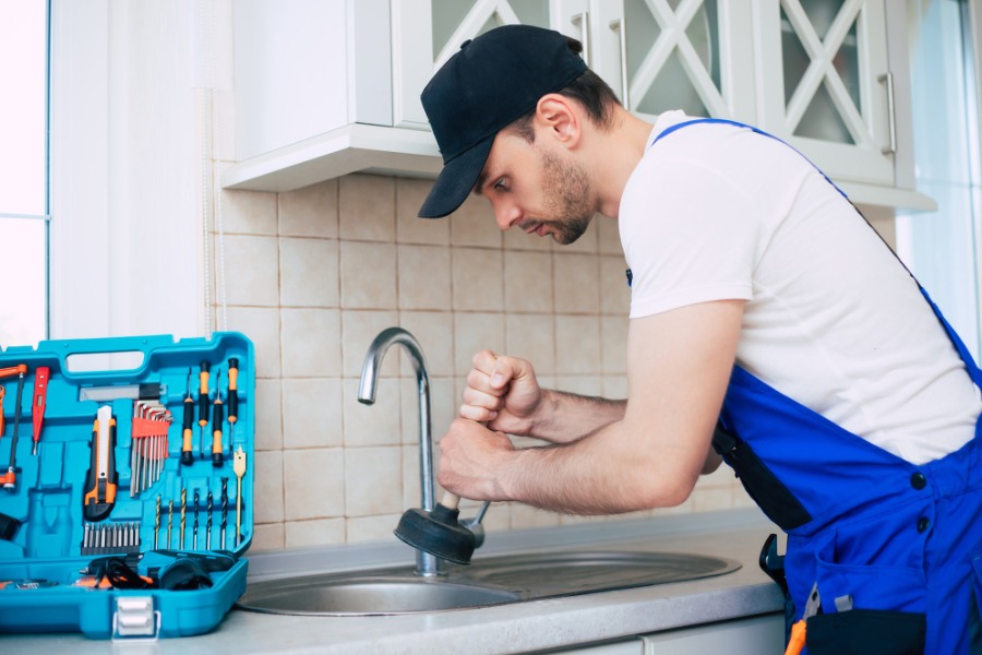 A man in a hat and blue overalls working on a kitchen sink to access the drain in order to clean it.