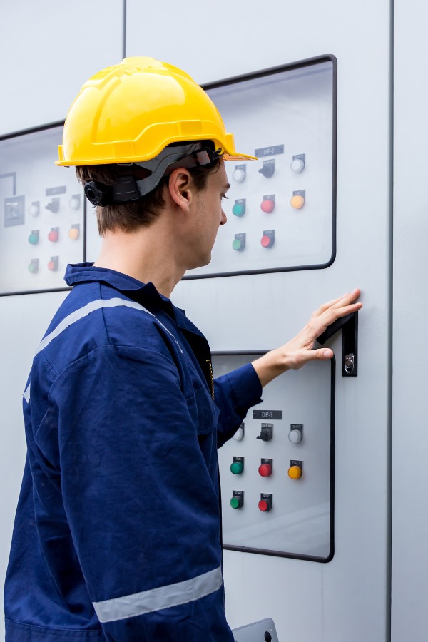 A man in a yellow hard hat and blue work outfit, at a large, commercial electrical panel.