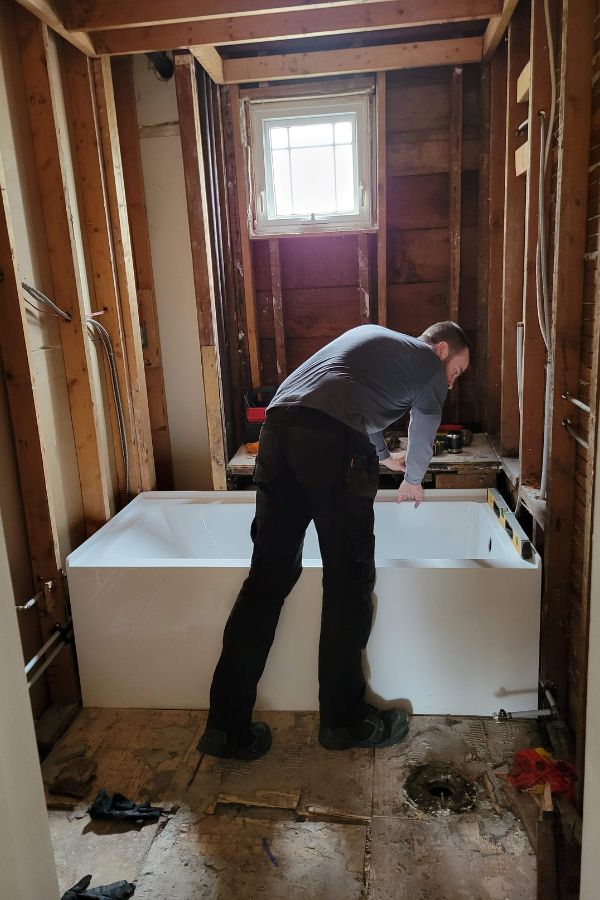 A man in a bathroom with the walls down to the studs, standing over a white alcove bathtub.
