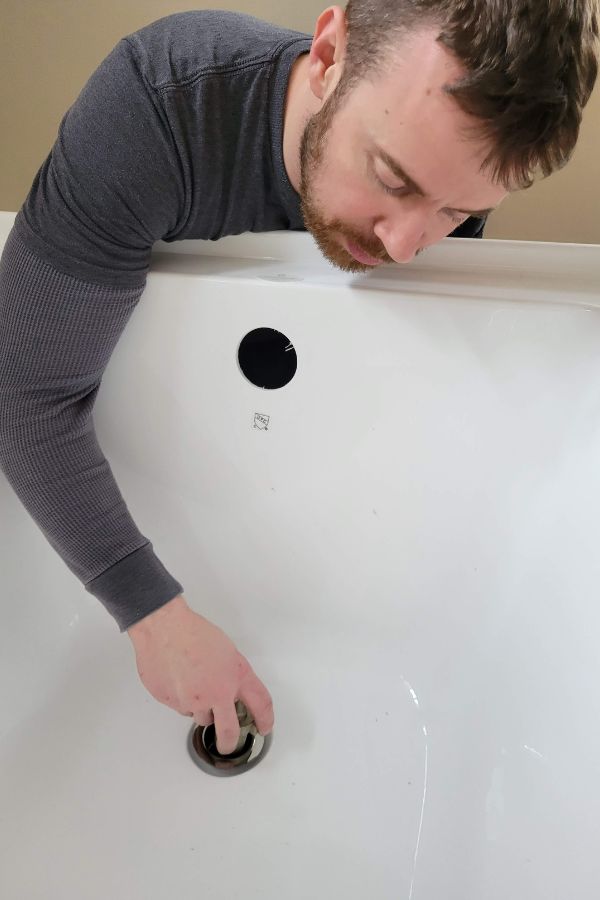 A man installing a white bathtub with his arm inside of a drain.